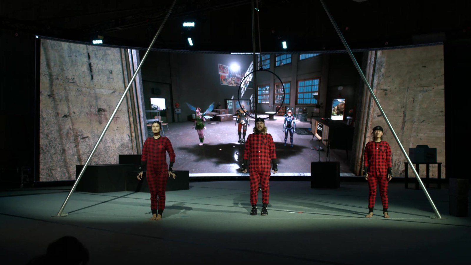 What the in-venue audience saw — the three circus performers in red plaid pyjama costumes (hiding active-marker mocap suits underneath, with passive markers on the hats and gloves for head and hand tracking — a hybrid mocap setup) standing on stage in front of the wraparound LED wall, with the live virtual render visible behind them showing the three characters they're driving (fairy on the left, Wukong centre, silver-and-horned juggler on the right), in matching positions. A 1:1 mapping between physical performers and digital characters, shared by everyone in the room.